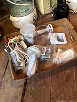Close-up of various mixer attachments in cardboard box including beaters, whisks, and plastic parts along with power cord.
