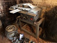 Ryobi 10in BT3000 table saw with gray metal stand. Various cables and parts on the floor.