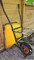 Side view of yellow wheelbarrow showing black metal frame with rubber handle grips and single wheel with visible rust.