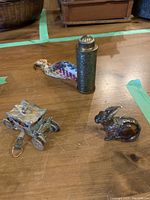 Photo showing three antique sewing items on a wooden surface, including a metal carriage dispenser, a bunny-shaped dispenser, and the cylindrical tape measure dispenser with colorful woven tape.