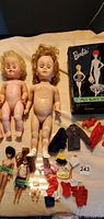 Photo showing three large nude dolls labeled as 1958 Barbie, 1958 Midge, and 1963 Skipper with various doll clothes and black 1962 Barbie travel case.