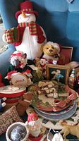 Large stuffed snowman, teddy bears, Christmas plates, framed needlepoint, ornaments, and small decorations shown together to display variety of lot contents.