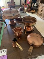 Large copper food warmer with lid and stand, small pot, pan with wooden handle, and small coffee pot on table.