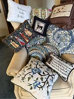 Assorted decorative pillows arranged on a beige upholstered chair, showing various embroidered and needlepoint designs including floral, animal, and text motifs, along with two braided round placemats.