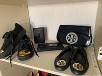 First shelf: black leather sneakers with gold toe cap, black card holder embossed star, Tory Burch ridged wallet, Tory Burch envelope clutch, Tory Burch Reva flats