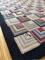 Close-up image showing the geometric square pattern in multi-colors with a black border along the edge of the rug. The loops of the yarn material are visible.