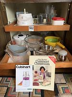 Overview of kitchen items including Cuisinart food processor, glass measuring cups, strainers, colanders, and cookbooks in open cabinet drawer