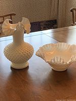 Photo showing both the milk glass fluted hobnail vase and bowl on a wooden table with chairs in background.