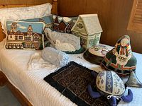 Photo of the entire collection of needlepoint pillows and door weights arranged on a bed, showing various sizes and designs including house scenes, a lamb pillow, and assorted door weights shaped like animals and figures.
