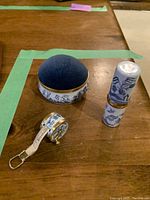 Photo showing all three pieces: pincushion, measuring tape, and needle holder with blue fabric, blue and white designs, and gold trim.