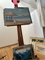 Wide view of wooden easel holding a beach landscape painting and additional canvas boards and framed art leaning against the wall on the floor.
