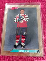 Brett Hull hockey trading card on a red textured surface, showing front artwork with player in uniform and gold border.