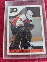 Photo of Pelle Lindbergh hockey card showing player in white goalie mask and Flyers uniform in protective plastic case.
