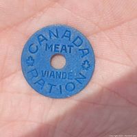 Close-up of a blue circular meat ration token held in palm of hand. Token has a central hole and embossed wording 'CANADA MEAT RATION VIANDE'.