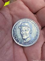 Hand-held photo of aluminum baseball token coin featuring Jackie Robinson with clear view of portrait and inscription.