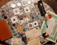 Overview of coins, stamps, jewelry, documents, and collectibles spread on marble table.