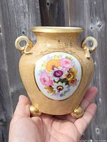 Front view of vase held in hand showing detailed floral painting and gold finish