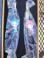 Two metal sticks with figurine toppers in blue and pink, sealed in plastic inside a black box.