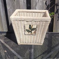Front view of a rectangular wicker bathroom waste basket showing natural wicker weave with a decorative green leaf inset.
