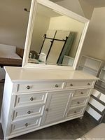 Front view of white dresser with 8 drawers and central cabinet with a large white framed mirror on top.