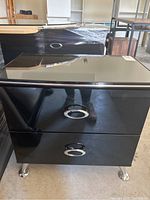 Front view of a black lacquer bedside table showing the two drawers with chrome oval handles and metal legs, placed in a warehouse.
