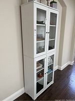Front view of tall white display cabinet with six glass shelves behind double doors, containing various items.
