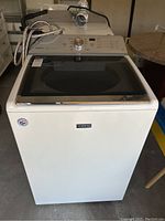 Front view of Maytag Bravos XL top-loading washer showing white exterior, glass lid, and hoses on top