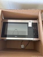 Exterior view of Whirlpool microwave with stainless steel face and black control panel, showing power cord inside box