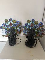 Front view of two stained glass peacock lamps with multi-colored fan-shaped shades and metal peacock bases.
