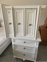 Photo showing two white bedside chests stacked, each with three paneled drawers and silver knobs, reflecting some light from surface.