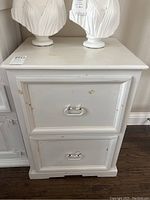 Front view of one white wooden file cabinet showing two drawers with white metal drawer pulls. Paint chips and scratches visible on drawer fronts and cabinet top.