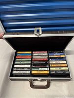 Open hard-sided brown travel case displaying 31 assorted vintage cassette tapes arranged in three rows.