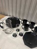 Photo displaying MCM black handled glass pitcher with stir stick, three small matching clear glass cups with black bases, stacked black octagonal dinner plates, side plates, and black mugs from Arcoroc France Octime Black pattern.