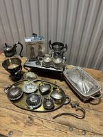 Overview photo of the lot items arranged on wooden surface showing multiple silver plate serving pieces, trinket boxes, and silver plate and glass carafe with stand and burner.