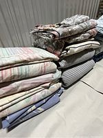 Multiple folded vintage bed sheet sets stacked showing pastel stripes, floral, and neutral colored fabrics
