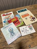 Front view of six vintage cartoon books stacked and spread on wooden table, showing colorful cartoon covers including Herman Treasury, Marmaduke, Family Circus, Saxtons World of Business, and Crazy World of Golf.