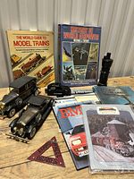 Photo showing vintage model cars, brake sign, multiple books including model train guide, and magazines on trains and railways, plus a small model locomotive. Items laid out on table.