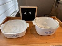 View of both Corningware casserole dishes with lids on a wooden table with a sign labeled LOT 42 in the background.