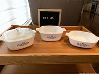 Three blue cornflower Corningware casserole dishes displayed with one clear glass lid fitting the left dish.