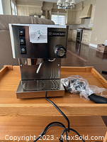 Front view of stainless steel Rancilio Silvia machine on wooden table, steam wand extended and drip tray in place, plastic-wrapped portafilter beside it