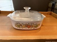 White Corningware casserole dish with Spice of Life vegetable pattern and clear glass lid, photographed on wood tray.