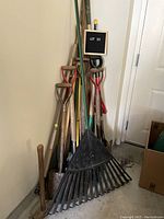 Overview photo of large garden tool lot including various shovels, rakes, hoes, and other long-handled garden tools leaning against a wall.