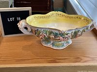 Side view of antique Italian footed bowl featuring sculpted cherubs and Father Wind face handles, placed on wooden surface with black lot sign visible.