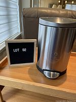 Mini stainless steel garbage can with black lid and foot pedal beside a sign reading LOT 92, set on a wooden table with a window and furniture in the background.