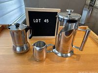 Photo showing all three items: stainless steel coffee press pot with plunger, creamer, and frothing cup, placed on a wooden table with lot number board.