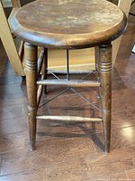 Vintage wooden stool showing round seat, four tapered legs with decorative turnings, and horizontal stretchers. Wear marks and patina visible.