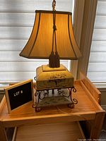 Front view of the vintage floral tin table lamp with fabric shade and tassel pull chain, showing detailed floral painting on base and lower shelf.