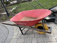 Side angled view of the red metal wheelbarrow showing wooden handles and yellow wheel on paved ground.