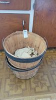 Two wooden 2 bushel baskets stacked, showing black metal support bands, wooden slats, and wire handles, with one containing a cloth.