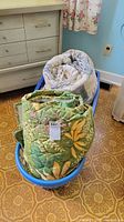 Photo showing two rolled quilted blankets, one green floral and one light geometric pattern, placed inside a blue plastic bin.